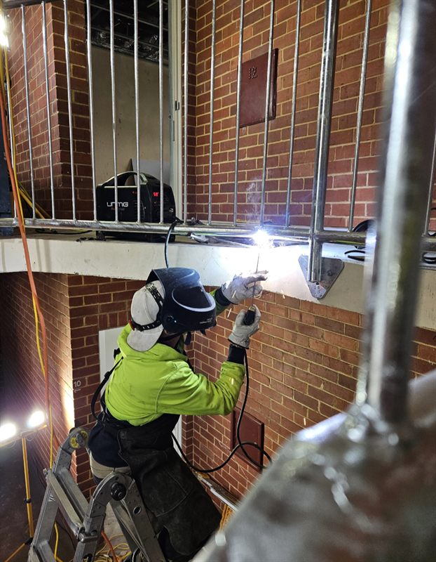 Welder in protective clothing welding a metal railing beside a brick wall